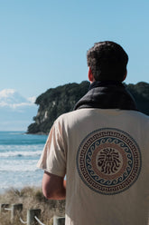 A person facing away from the camera stands by the sea, wearing a beige t-shirt with a circular emblem design on the back.